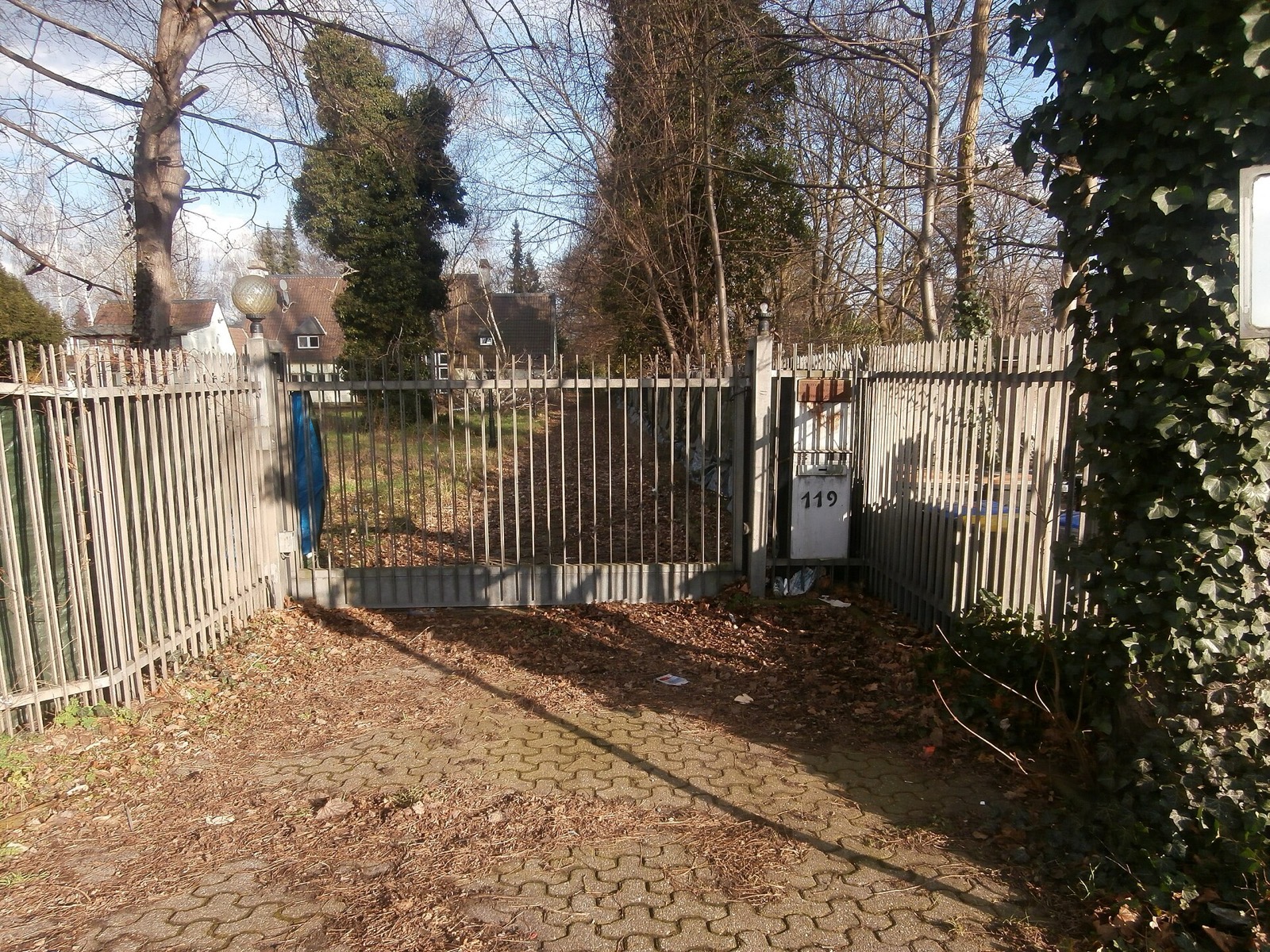 Verschlossenes Metalltor an der Bonner Landstraße in Köln-Hahnwald, dahinter ein großzügiges Grundstück mit altem Baumbestand