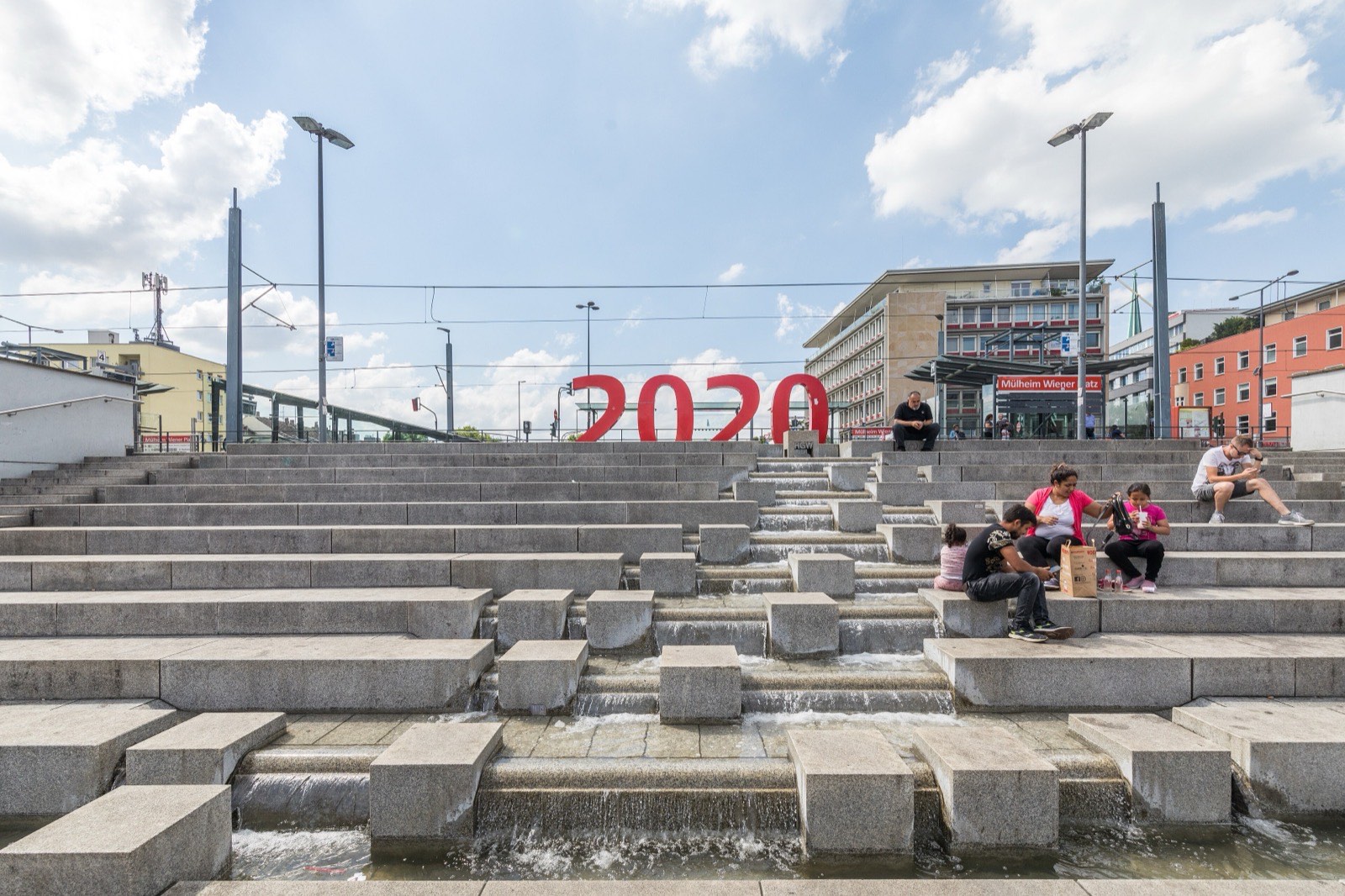 Die Wassertreppe am Wiener Platz in Köln-Mülheim, auf den Stufen sitzen Menschen in der Sonne