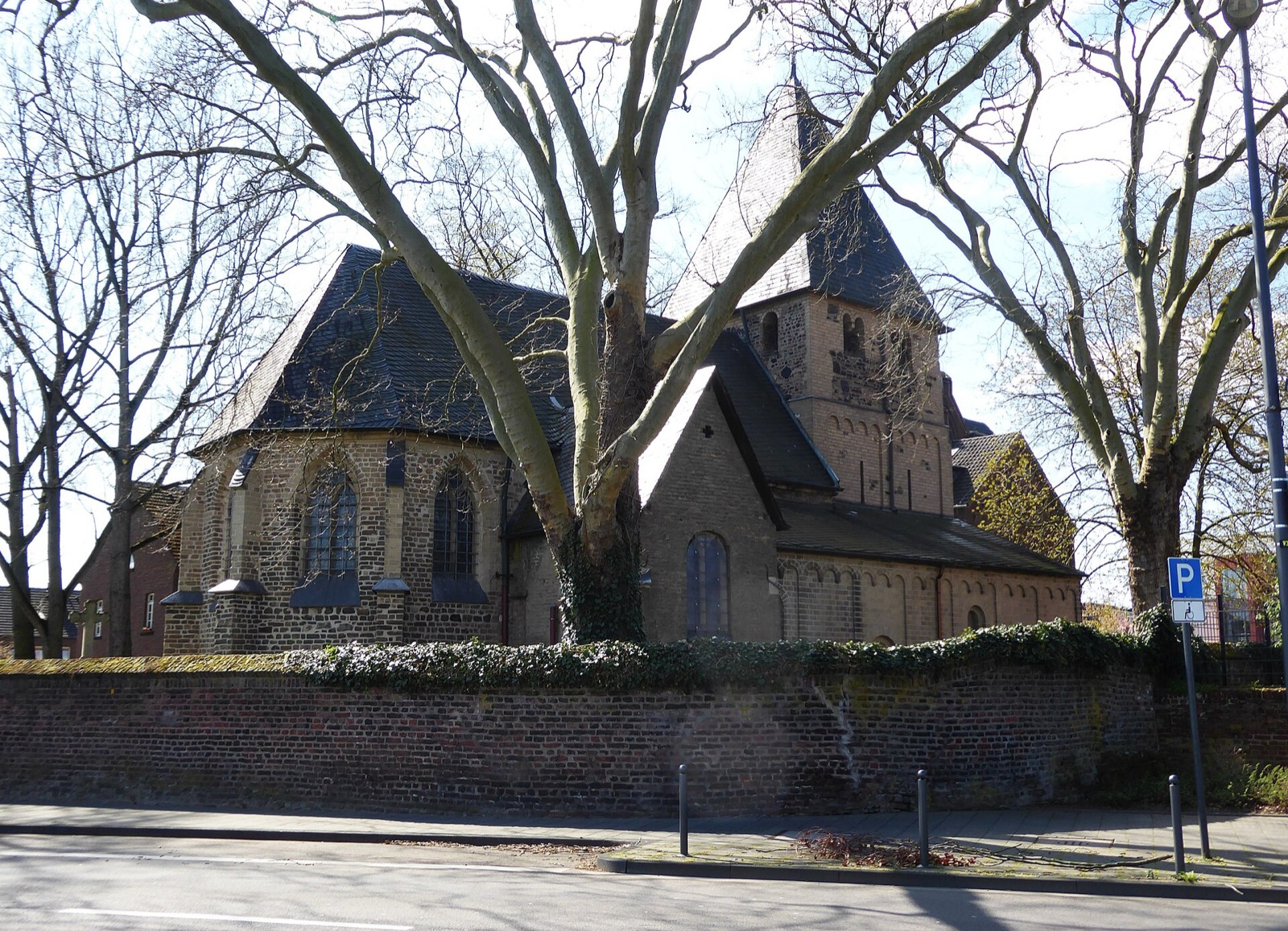 Alt St. Katharina in Köln-Niehl: mittelalterliche Kirche aus Bruchstein hinter alten Platanen und einer Backsteinmauer