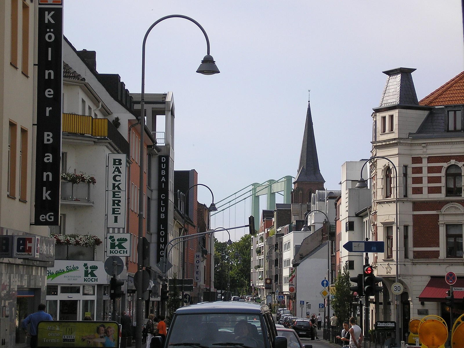 Die Hauptstraße in Köln-Rodenkirchen mit Geschäften im Erdgeschoss, Gründerzeithäusern und dem Kirchturm sowie der Rodenkirchener Autobahnbrücke im Hintergrund