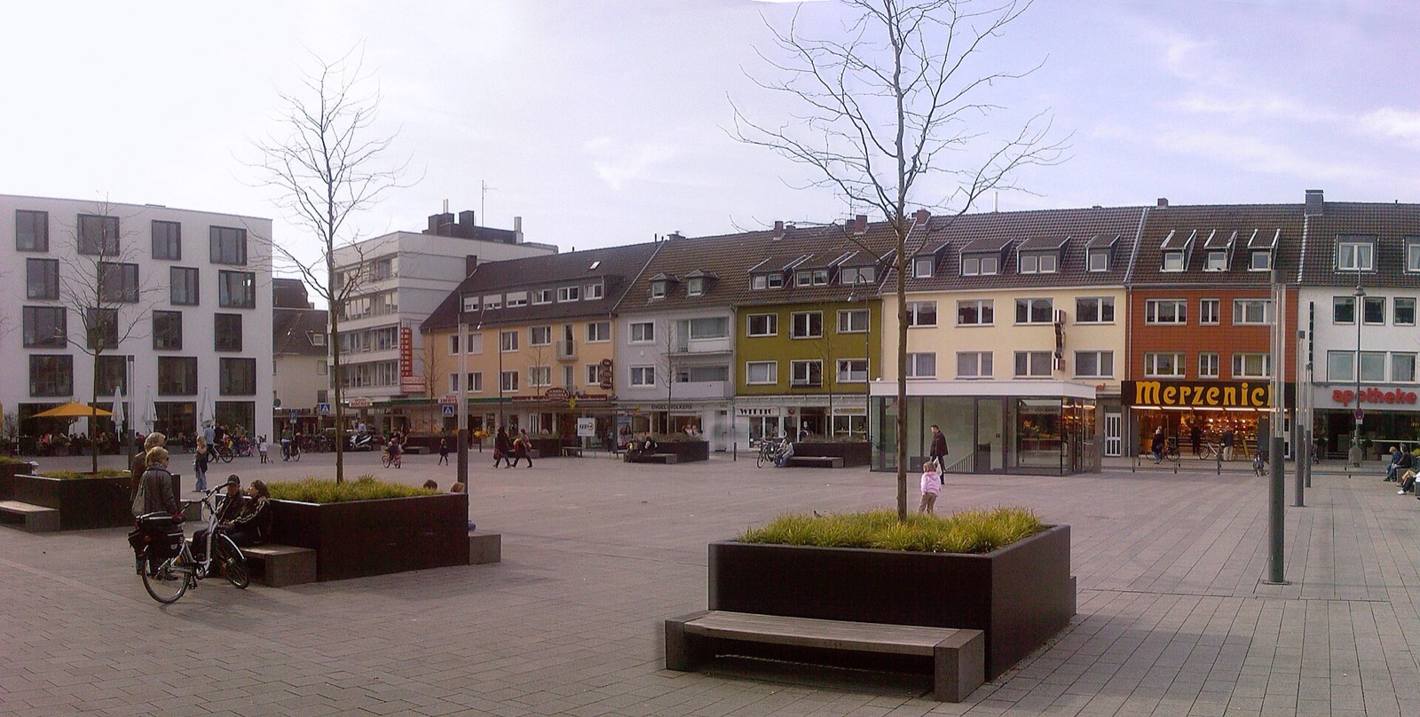 Der Maternusplatz in Köln-Rodenkirchen mit gepflastertem Platz, bepflanzten Sitzelementen und Wohnhäusern mit Läden und Apotheke im Erdgeschoss
