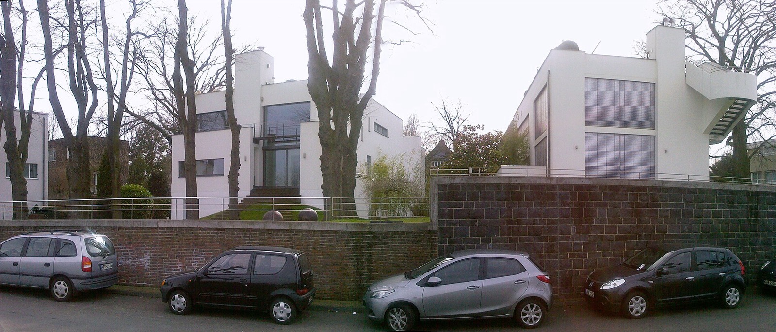 Zwei weiße Wohnhäuser im Stil der klassischen Moderne an der Uferstraße in Köln-Rodenkirchen, oberhalb einer Bruchsteinmauer zwischen alten Platanen