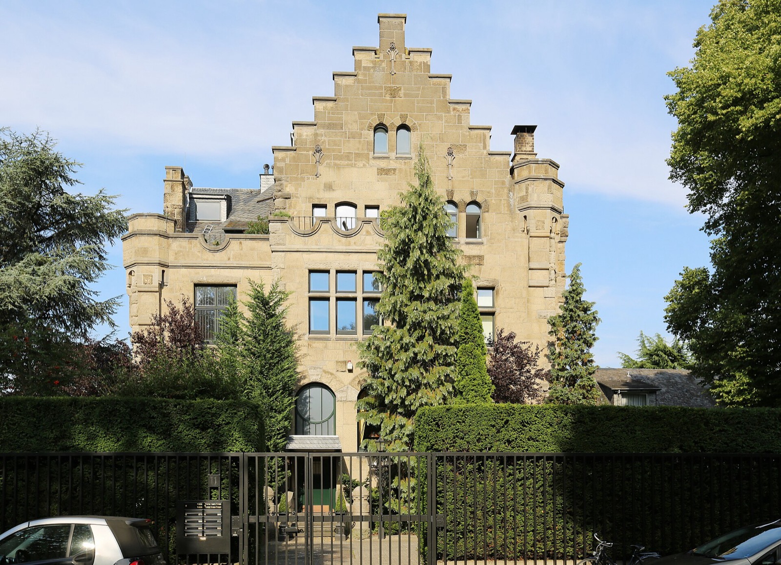 Villa Malta an der Hauptstraße 11 in Köln-Rodenkirchen: burgähnliche Natursteinvilla mit Staffelgiebel und Ecktürmchen hinter Hecke und schmiedeeisernem Zaun